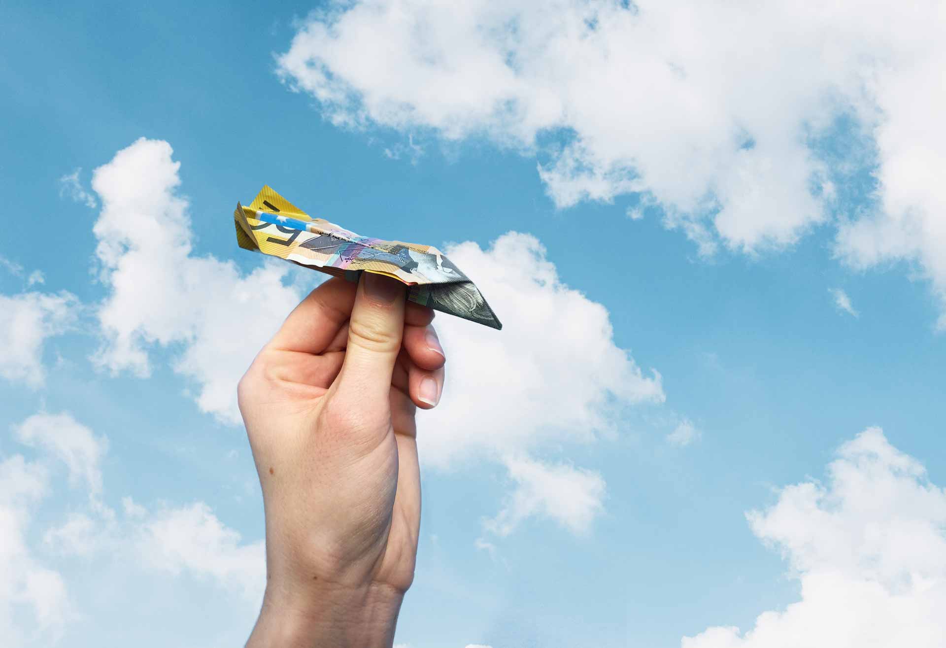 A hand holding an AU$50 banknote paper aeroplane against a blue sky